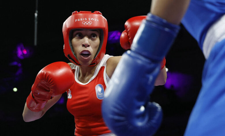 Uma boxeadora está em ação, usando um capacete vermelho e luvas vermelhas, enquanto se prepara para lutar. Ela está em um ringue de boxe, com um fundo desfocado que sugere um ambiente de competição. A adversária, visível à direita, está usando luvas azuis. A boxeadora parece concentrada e determinada, com uma expressão facial intensa.