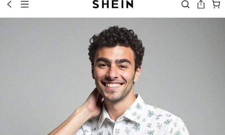 Um jovem sorridente está posando para a foto, usando uma camisa de botão branca com estampas de folhas verdes. Ele tem cabelo cacheado e está com uma mão na nuca, em um fundo cinza claro. A camisa é de manga curta e ele está usando calças escuras.