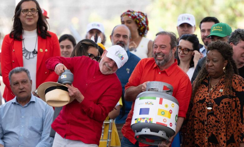 A imagem mostra um evento ao ar livre com várias pessoas reunidas. No centro, um homem de camisa vermelha e chapéu branco segura um botijão de gás, enquanto outro homem ao seu lado, vestido de azul, segura um recipiente semelhante. Ao fundo, há uma multidão de pessoas, algumas usando bonés e chapéus coloridos. O ambiente parece festivo, com sorrisos e interações entre os participantes.