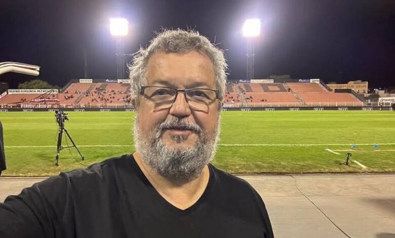 Um homem com barba grisalha e óculos está tirando uma selfie em um estádio de futebol à noite. Ao fundo, vê-se o campo iluminado por refletores e algumas arquibancadas com poucas pessoas. O homem está vestindo uma camiseta preta e parece estar em um ambiente descontraído.