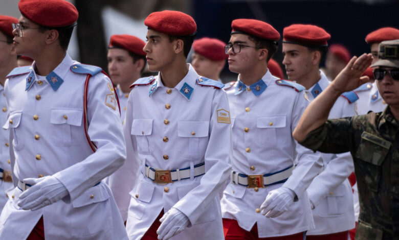 Um grupo de jovens soldados em uniforme branco e calças vermelhas, usando boinas vermelhas, marcha em formação. Eles estão em um desfile militar, com expressões sérias e focadas. Ao fundo, um soldado em uniforme camuflado está em posição de saudação.