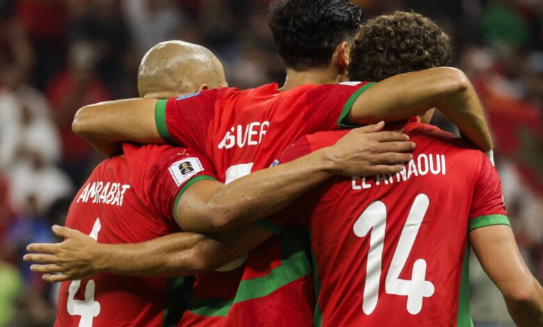 Três jogadores de futebol de Marrocos estão se abraçando em um momento de comemoração. Eles vestem uniformes vermelhos com detalhes verdes. O jogador à esquerda tem o número 4 nas costas, o do meio tem o número 7 e o da direita tem o número 14. O fundo mostra uma multidão, sugerindo um ambiente de estádio.