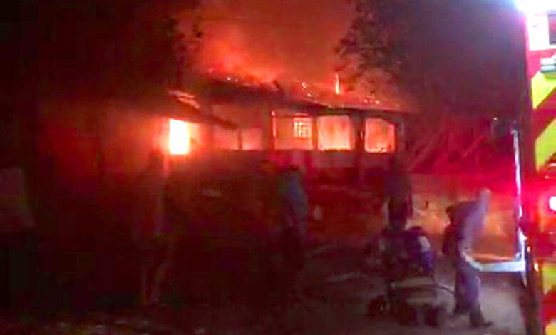 A imagem mostra uma estrutura em chamas durante a noite, com fumaça e fogo visíveis. Há pessoas próximas à cena, possivelmente tentando controlar o incêndio. Um veículo de emergência, possivelmente um caminhão de bombeiros, está presente à direita, iluminando a área com suas luzes.