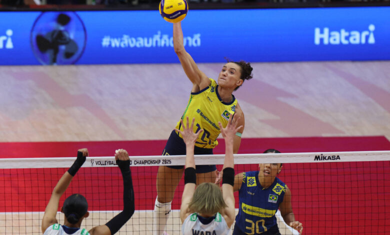 Uma jogadora de vôlei está saltando para fazer um ataque, segurando uma bola amarela. Ela está vestindo um uniforme amarelo e azul, enquanto duas jogadoras adversárias, uma com a camisa número 15 e outra com a número 13, estão posicionadas na rede. O fundo mostra uma quadra de vôlei com uma bandeira e um painel publicitário.