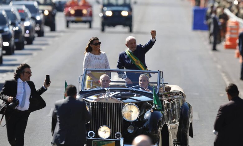 Uma cena de um desfile com um carro conversível. No carro, há um homem acenando e uma mulher ao seu lado. Eles estão cercados por pessoas em trajes formais, algumas tirando fotos. Ao fundo, há uma fila de veículos e pessoas assistindo ao evento. O carro é de cor escura e possui uma bandeira do Brasil.