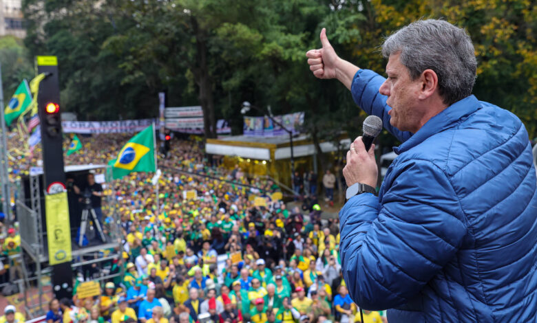A imagem mostra uma grande multidão em uma manifestação ao ar livre. No primeiro plano, um homem com cabelo grisalho e vestido com uma jaqueta azul está falando ao microfone e fazendo um gesto de positivo com a mão. Ao fundo, há várias bandeiras do Brasil e pessoas vestindo roupas nas cores da bandeira. A cena é em um ambiente arborizado, com árvores ao redor e um clima nublado.