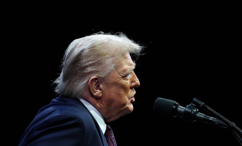 A imagem mostra um homem de perfil, com cabelo loiro e uma jaqueta escura, falando em um microfone. O fundo é escuro, destacando a figura do homem que parece estar em um evento ou discurso.