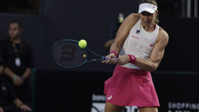 Uma jogadora de tênis está em ação, prestes a rebater uma bola amarela com sua raquete. Ela usa uma blusa branca e uma saia rosa, com uma faixa na cabeça. Ao fundo, é possível ver algumas pessoas assistindo ao jogo, em um ambiente de quadra de tênis.