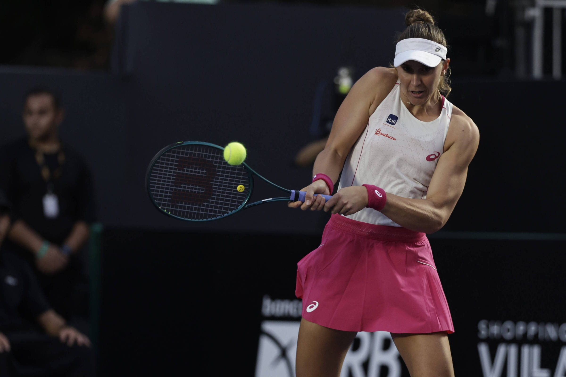 Uma jogadora de tênis está em ação, prestes a rebater uma bola amarela com sua raquete. Ela usa uma blusa branca e uma saia rosa, com uma faixa na cabeça. Ao fundo, é possível ver algumas pessoas assistindo ao jogo, em um ambiente de quadra de tênis.