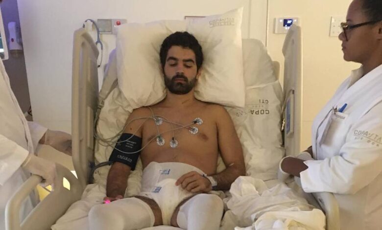 Um homem está deitado em uma cama hospitalar, com o tronco nu e conectado a monitores cardíacos. Ele tem um olhar sério e está cercado por duas profissionais de saúde, uma delas segurando um equipamento. O ambiente é claro e limpo, com paredes brancas e um pouco de equipamento médico visível ao fundo.