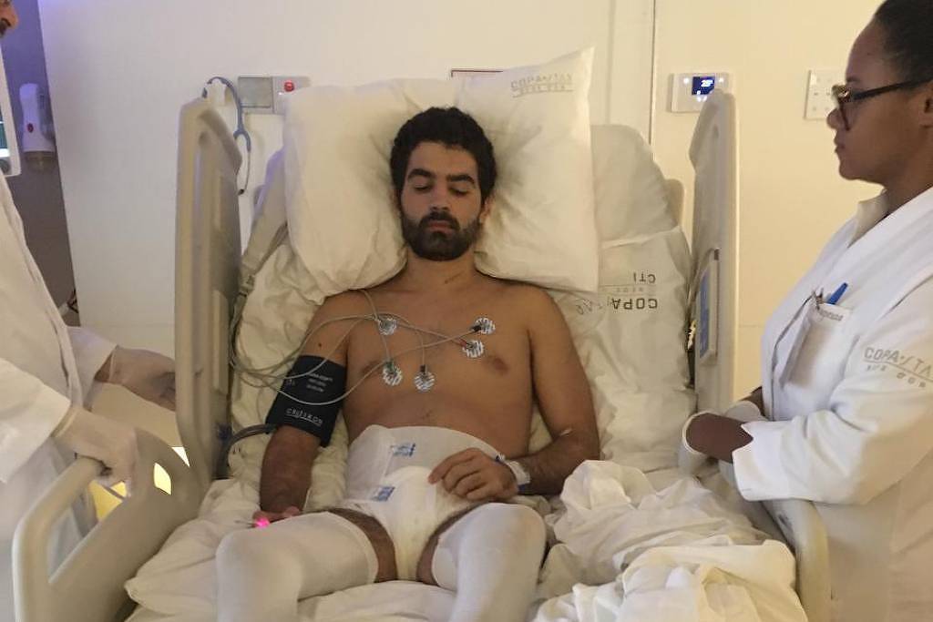 Um homem está deitado em uma cama hospitalar, com o tronco nu e conectado a monitores cardíacos. Ele tem um olhar sério e está cercado por duas profissionais de saúde, uma delas segurando um equipamento. O ambiente é claro e limpo, com paredes brancas e um pouco de equipamento médico visível ao fundo.