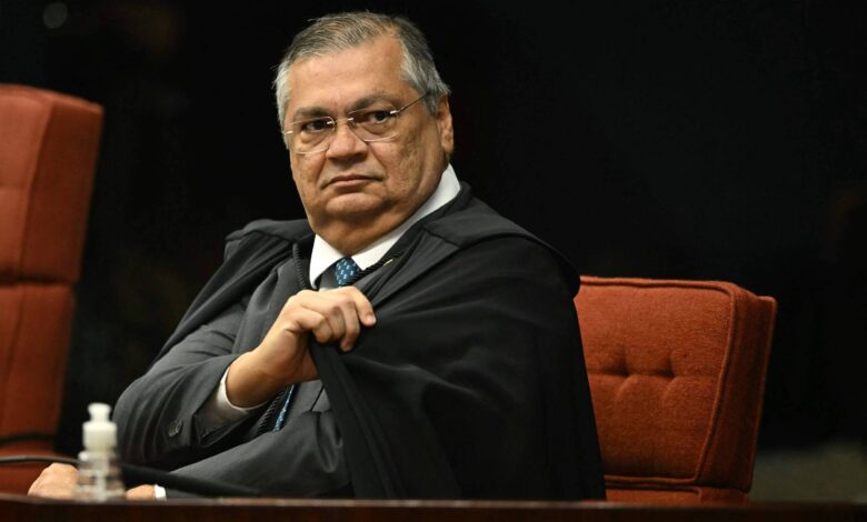 Um homem de meia-idade, com cabelo grisalho e óculos, está sentado em uma cadeira em um ambiente formal. Ele usa uma toga preta e uma camisa branca com uma gravata azul. O homem parece estar prestando atenção ou avaliando a situação ao seu redor. Ao lado dele, há uma garrafinha de água sobre a mesa. O fundo é escuro, com cadeiras vermelhas visíveis.