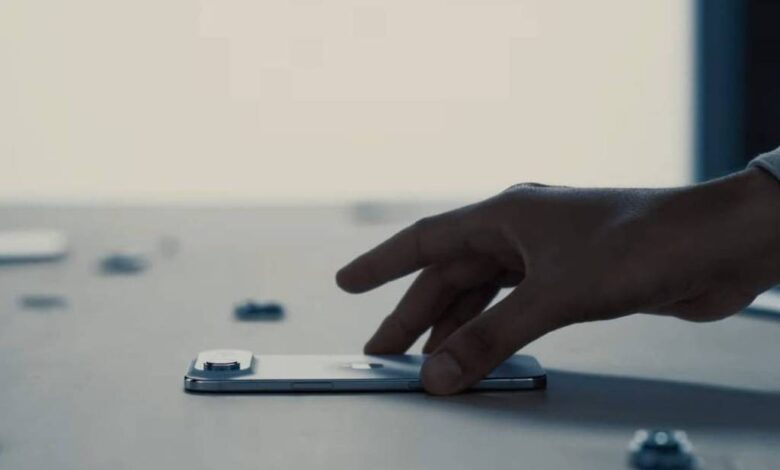 A imagem mostra uma mão humana posicionada sobre um smartphone em uma mesa. Ao redor do smartphone, há vários pequenos objetos que parecem ser chips ou cartões. O fundo é desfocado, com uma iluminação suave, sugerindo um ambiente de trabalho ou uma configuração minimalista.