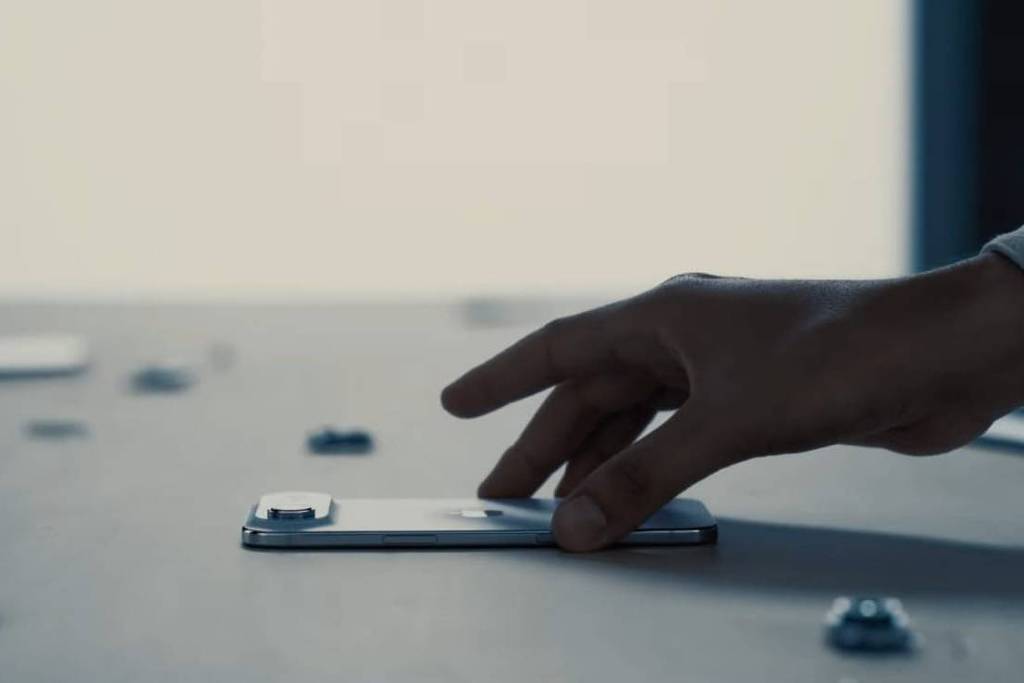 A imagem mostra uma mão humana posicionada sobre um smartphone em uma mesa. Ao redor do smartphone, há vários pequenos objetos que parecem ser chips ou cartões. O fundo é desfocado, com uma iluminação suave, sugerindo um ambiente de trabalho ou uma configuração minimalista.