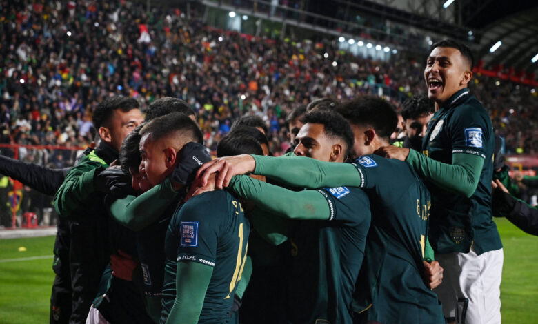 Um grupo de jogadores de futebol está se abraçando em celebração após um gol. Eles estão vestindo uniformes verdes e estão em um estádio cheio de torcedores. O ambiente é festivo, com muitos fãs ao fundo, alguns segurando bandeiras e outros demonstrando alegria. A iluminação do estádio é intensa, destacando a emoção do momento.