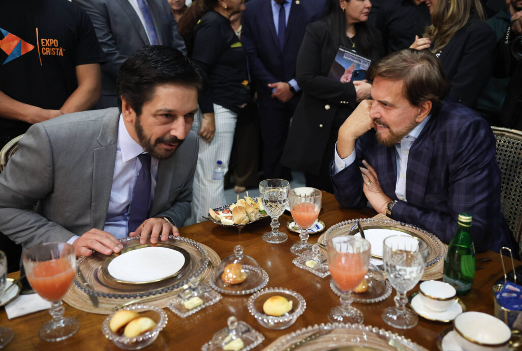 A imagem mostra dois homens sentados à mesa em um evento. Eles estão conversando, com pratos e copos à sua frente. Um dos homens está vestido com um terno cinza e uma camisa branca, enquanto o outro usa um terno azul escuro com uma camisa clara. Há bebidas em copos de cristal e pratos com doces sobre a mesa. Ao fundo, é possível ver outras pessoas e um ambiente de evento formal.