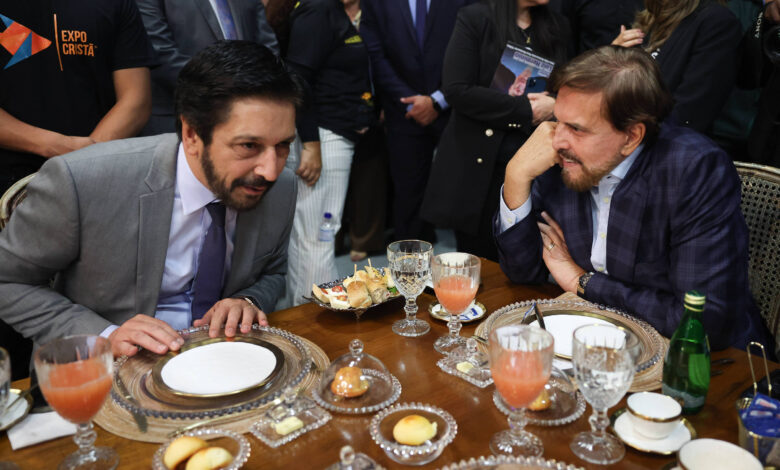 A imagem mostra dois homens sentados à mesa em um evento. Eles estão conversando, com pratos e copos à sua frente. Um dos homens está vestido com um terno cinza e uma camisa branca, enquanto o outro usa um terno azul escuro com uma camisa clara. Há bebidas em copos de cristal e pratos com doces sobre a mesa. Ao fundo, é possível ver outras pessoas e um ambiente de evento formal.