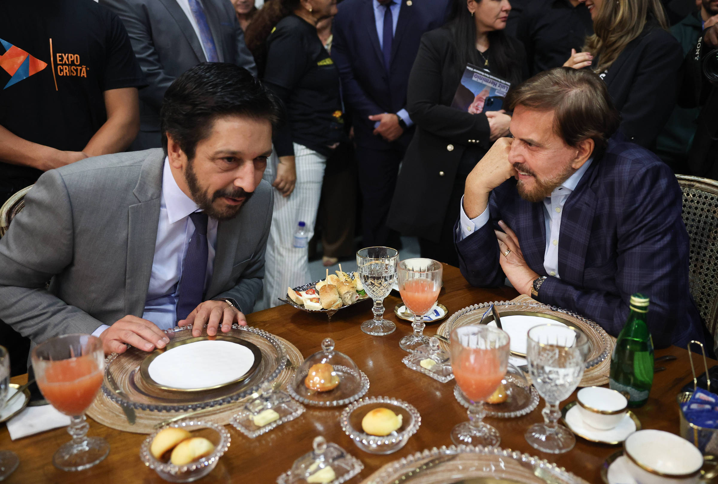 A imagem mostra dois homens sentados à mesa em um evento. Eles estão conversando, com pratos e copos à sua frente. Um dos homens está vestido com um terno cinza e uma camisa branca, enquanto o outro usa um terno azul escuro com uma camisa clara. Há bebidas em copos de cristal e pratos com doces sobre a mesa. Ao fundo, é possível ver outras pessoas e um ambiente de evento formal.
