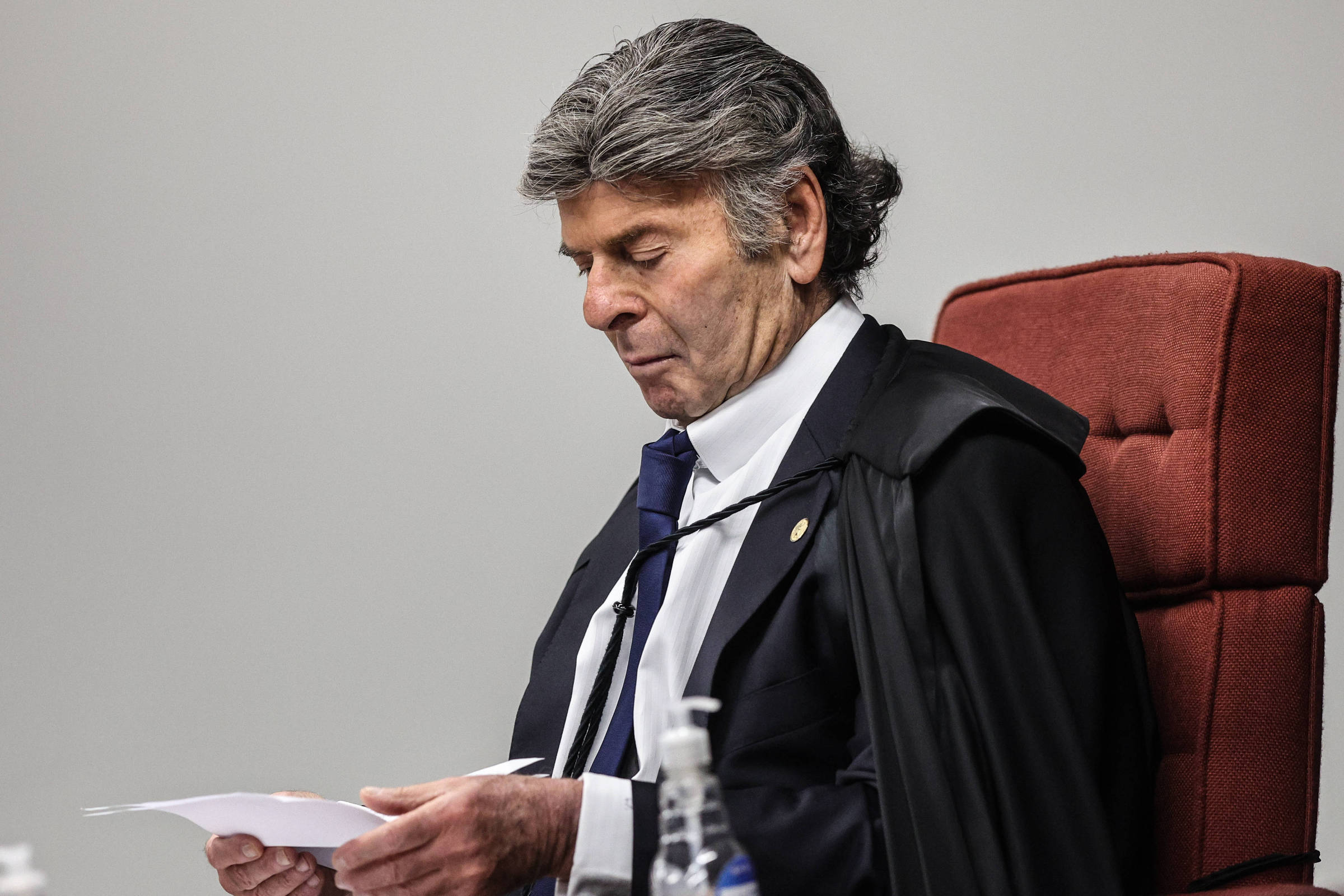 Um juiz está sentado em uma cadeira vermelha, com um manto preto. Ele está olhando para papéis que segura em suas mãos, com uma expressão séria. O ambiente parece ser uma sala de tribunal, com uma mesa e garrafas de água visíveis ao lado.