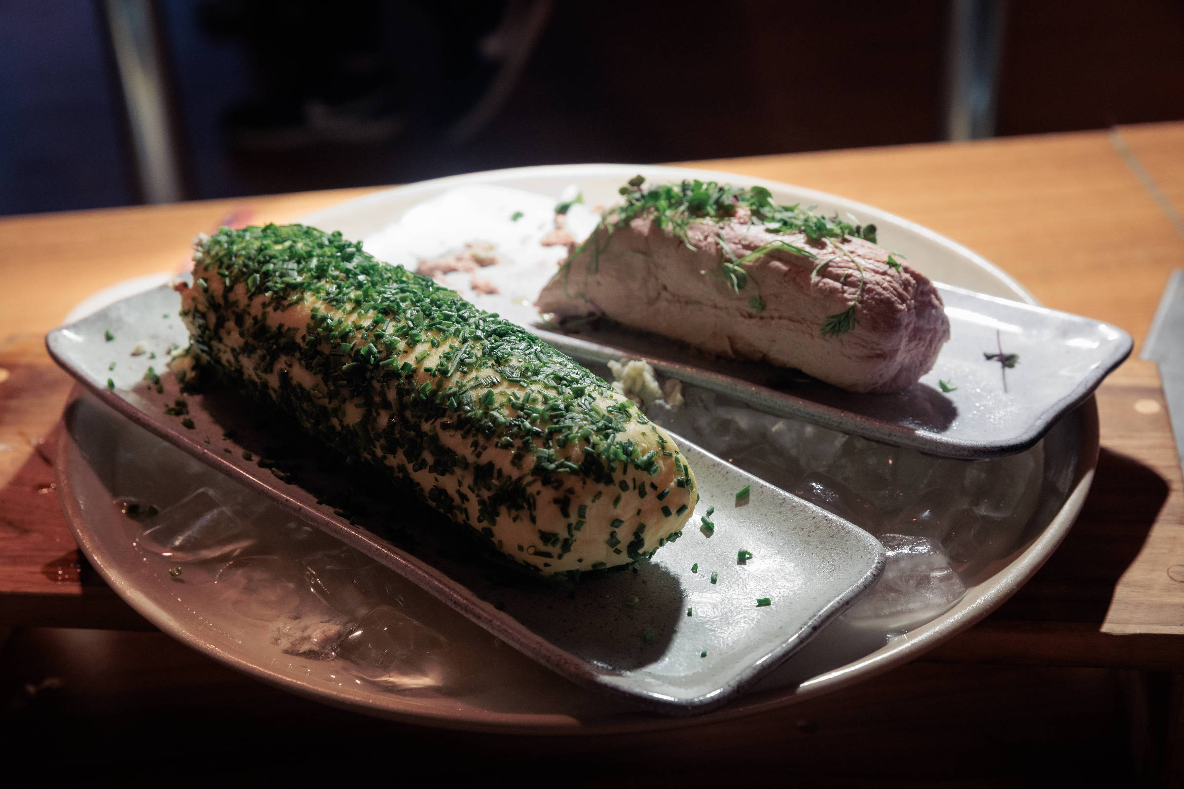 Um prato com duas porções de manteiga temperada. A porção à esquerda é coberta com ervas verdes picadas, enquanto a porção à direita é de uma manteiga mais clara, também com ervas. Ambas estão servidas em pratos retangulares sobre uma superfície de madeira.