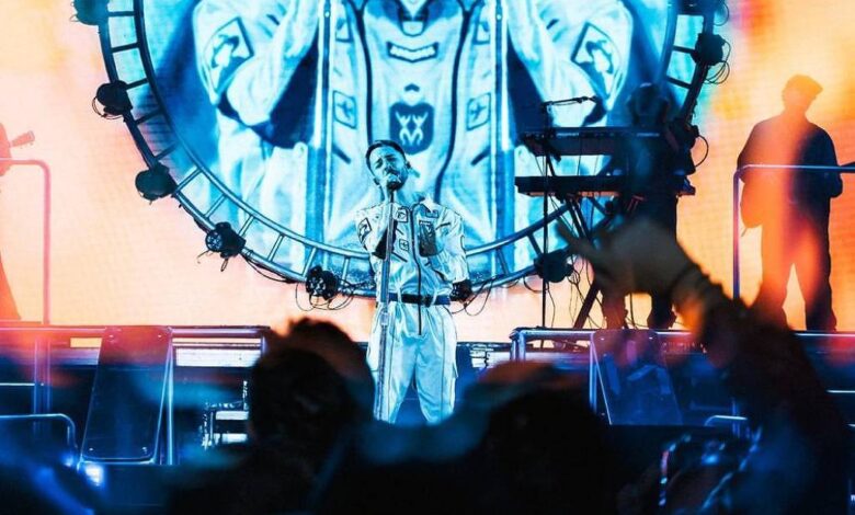 A imagem mostra um artista se apresentando em um palco iluminado com luzes coloridas. Ao fundo, há um grande painel circular que exibe uma imagem do artista em uma performance. O público é visível na parte inferior da imagem, com algumas pessoas levantando os braços. O ambiente é vibrante e cheio de energia, com uma mistura de luzes azuis e laranjas.