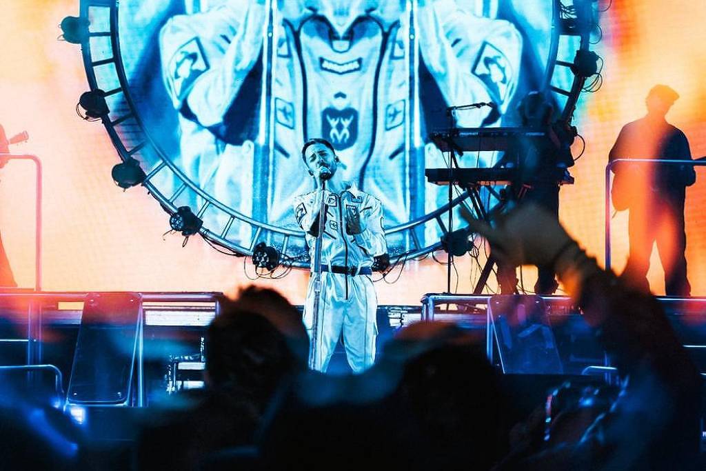 A imagem mostra um artista se apresentando em um palco iluminado com luzes coloridas. Ao fundo, há um grande painel circular que exibe uma imagem do artista em uma performance. O público é visível na parte inferior da imagem, com algumas pessoas levantando os braços. O ambiente é vibrante e cheio de energia, com uma mistura de luzes azuis e laranjas.