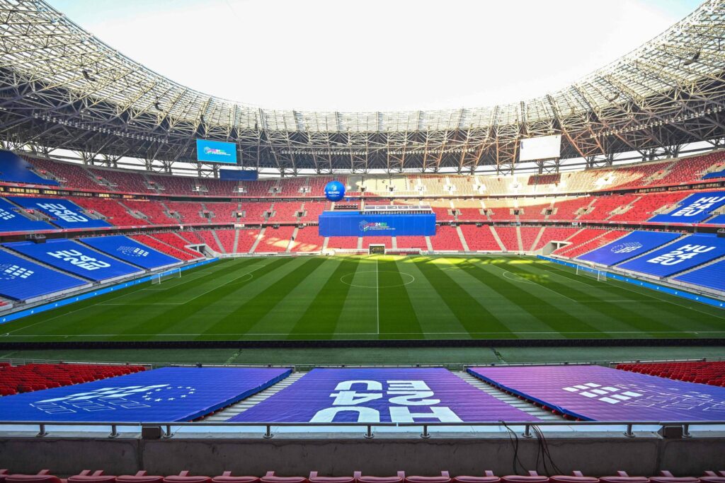 A imagem mostra um estádio de futebol vazio, com arquibancadas vermelhas e azuis. O campo está bem cuidado, com grama verde e listras visíveis. No fundo, há uma grande tela eletrônica e banners publicitários. A iluminação natural entra pelo teto do estádio, que é parcialmente coberto.