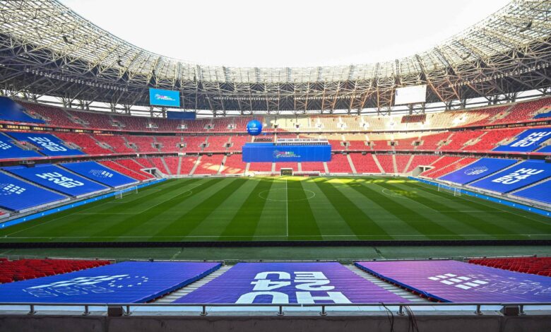 A imagem mostra um estádio de futebol vazio, com arquibancadas vermelhas e azuis. O campo está bem cuidado, com grama verde e listras visíveis. No fundo, há uma grande tela eletrônica e banners publicitários. A iluminação natural entra pelo teto do estádio, que é parcialmente coberto.