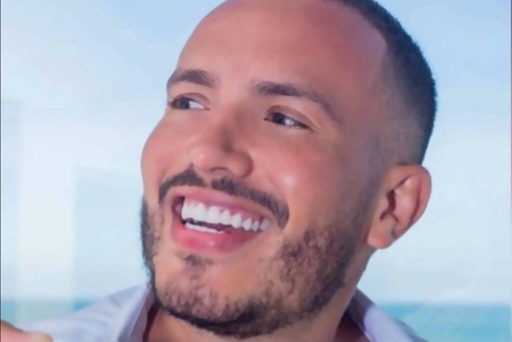 Um homem sorridente segurando uma xícara de café. Ele tem cabelo curto e barba bem aparada. O fundo mostra uma vista para o mar, sugerindo um ambiente relaxante. O homem está usando uma camisa branca aberta, revelando parte do peito.