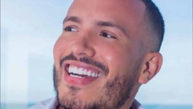 Um homem sorridente segurando uma xícara de café. Ele tem cabelo curto e barba bem aparada. O fundo mostra uma vista para o mar, sugerindo um ambiente relaxante. O homem está usando uma camisa branca aberta, revelando parte do peito.