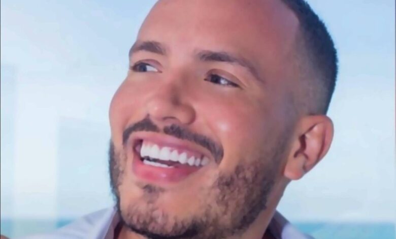 Um homem sorridente segurando uma xícara de café. Ele tem cabelo curto e barba bem aparada. O fundo mostra uma vista para o mar, sugerindo um ambiente relaxante. O homem está usando uma camisa branca aberta, revelando parte do peito.