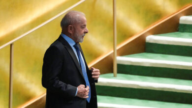 Um homem de terno escuro e gravata azul está em pé em um palco, com uma escada ao fundo. O homem tem cabelo grisalho e uma barba curta. O fundo é dourado e a escada é verde, com degraus cobertos de um material claro. O homem parece estar se preparando para falar ou se apresentar.
