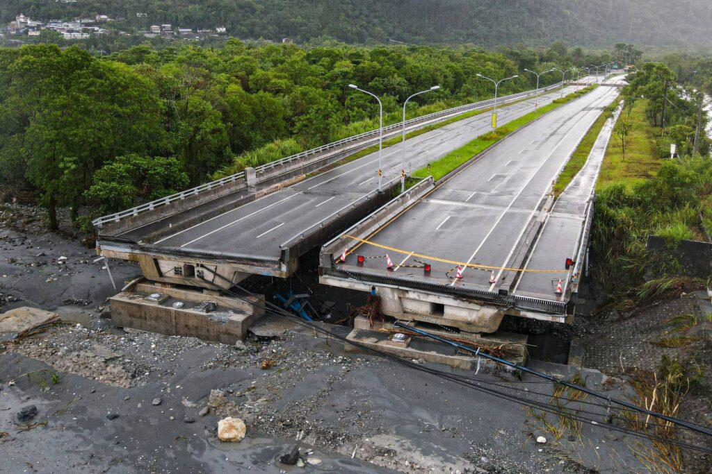 A imagem mostra uma ponte danificada, com uma parte dela colapsada e inclinada. O asfalto da estrada está visível, e há barreiras de segurança indicando que a passagem está bloqueada. Ao fundo, há uma vegetação densa e montanhas, sugerindo um ambiente natural. O céu está nublado, indicando condições climáticas possivelmente adversas.