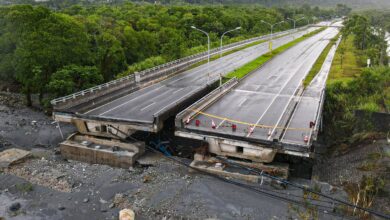A imagem mostra uma ponte danificada, com uma parte dela colapsada e inclinada. O asfalto da estrada está visível, e há barreiras de segurança indicando que a passagem está bloqueada. Ao fundo, há uma vegetação densa e montanhas, sugerindo um ambiente natural. O céu está nublado, indicando condições climáticas possivelmente adversas.
