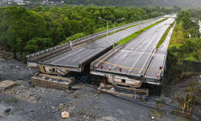 A imagem mostra uma ponte danificada, com uma parte dela colapsada e inclinada. O asfalto da estrada está visível, e há barreiras de segurança indicando que a passagem está bloqueada. Ao fundo, há uma vegetação densa e montanhas, sugerindo um ambiente natural. O céu está nublado, indicando condições climáticas possivelmente adversas.