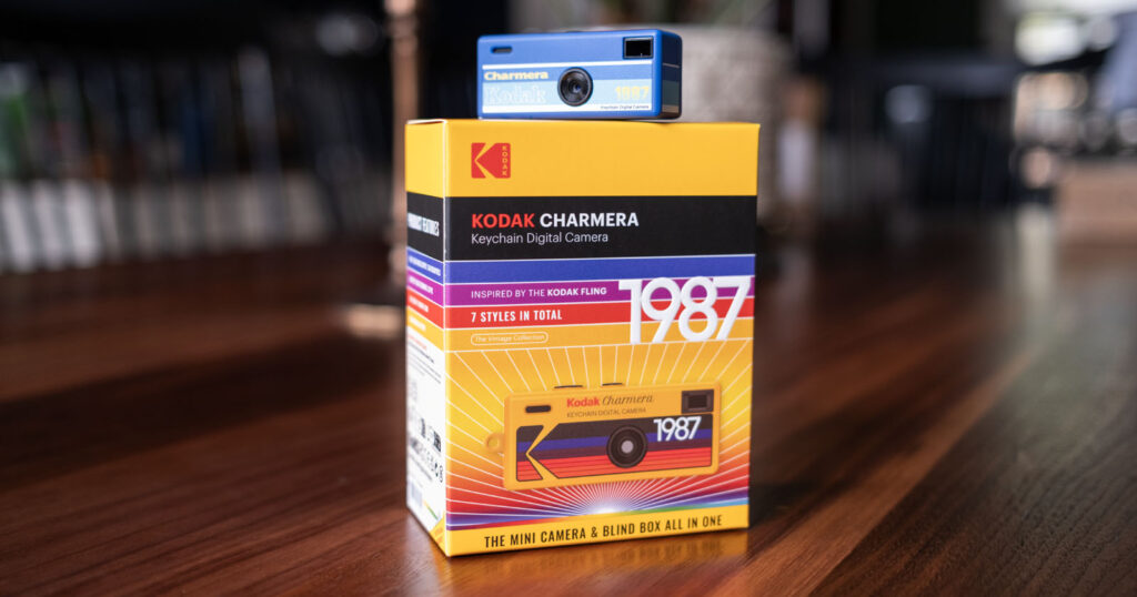 A blue Kodak Charmera keychain digital camera is placed on top of its colorful yellow and red box, which features bold