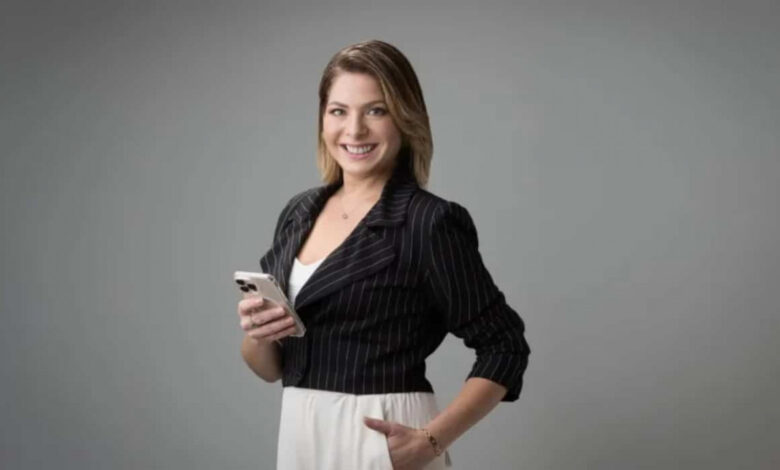 Daniela Lima de roupa formal branca e preta, sorrindo, com uma mão no bolso a e a outra segurando celular, olhando para a câmera