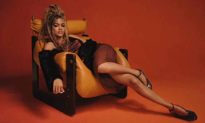Teyana Taylor lies in a plush light brown armchair wearing a sheer skirt