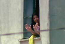 Mulher com cabelo preto aparece na janela aberta de um prédio claro, com as mãos levantadas em sinal de parada ou comunicação. Ela veste roupa clara e há um tecido amarelo pendurado no parapeito da janela.