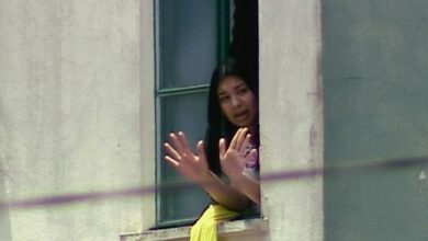 Mulher com cabelo preto aparece na janela aberta de um prédio claro, com as mãos levantadas em sinal de parada ou comunicação. Ela veste roupa clara e há um tecido amarelo pendurado no parapeito da janela.
