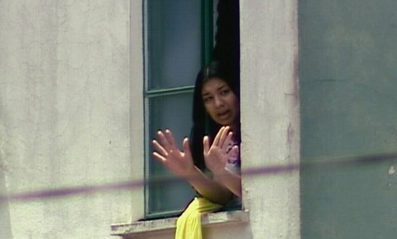 Mulher com cabelo preto aparece na janela aberta de um prédio claro, com as mãos levantadas em sinal de parada ou comunicação. Ela veste roupa clara e há um tecido amarelo pendurado no parapeito da janela.