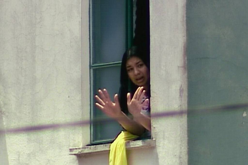 Mulher com cabelo preto aparece na janela aberta de um prédio claro, com as mãos levantadas em sinal de parada ou comunicação. Ela veste roupa clara e há um tecido amarelo pendurado no parapeito da janela.
